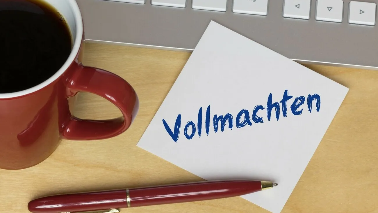 Ein Zettel mit der Aufschrift "Vollmachten" liegt halb auf einer Tastatur. Daneben steht eine rote Kaffeetasse und darunter ein roter Kugelschreiber.